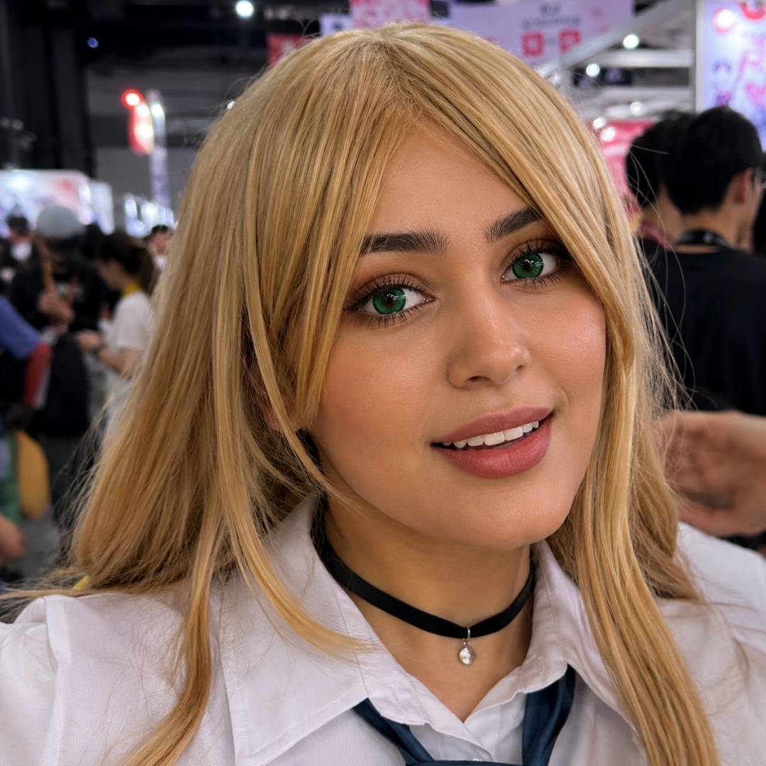Convention portrait of a woman with blonde hair wearing a school inspired cosplay outfit and black choker. Her smile and reem adult green contacts create a bright and playful appearance.
