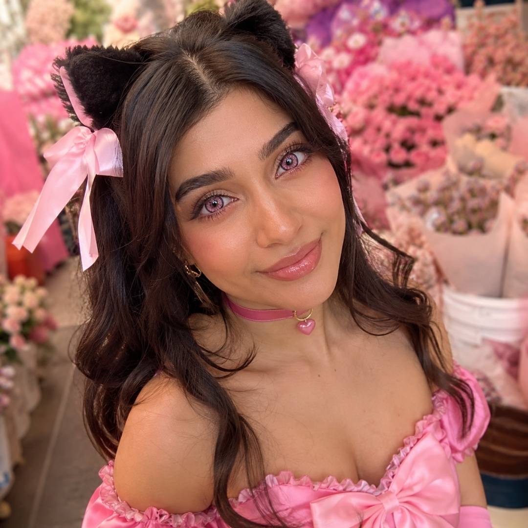 Anime-inspired cosplay with pink twintails, a ribbon choker, and a frilly pink dress, posing under cherry blossom trees.