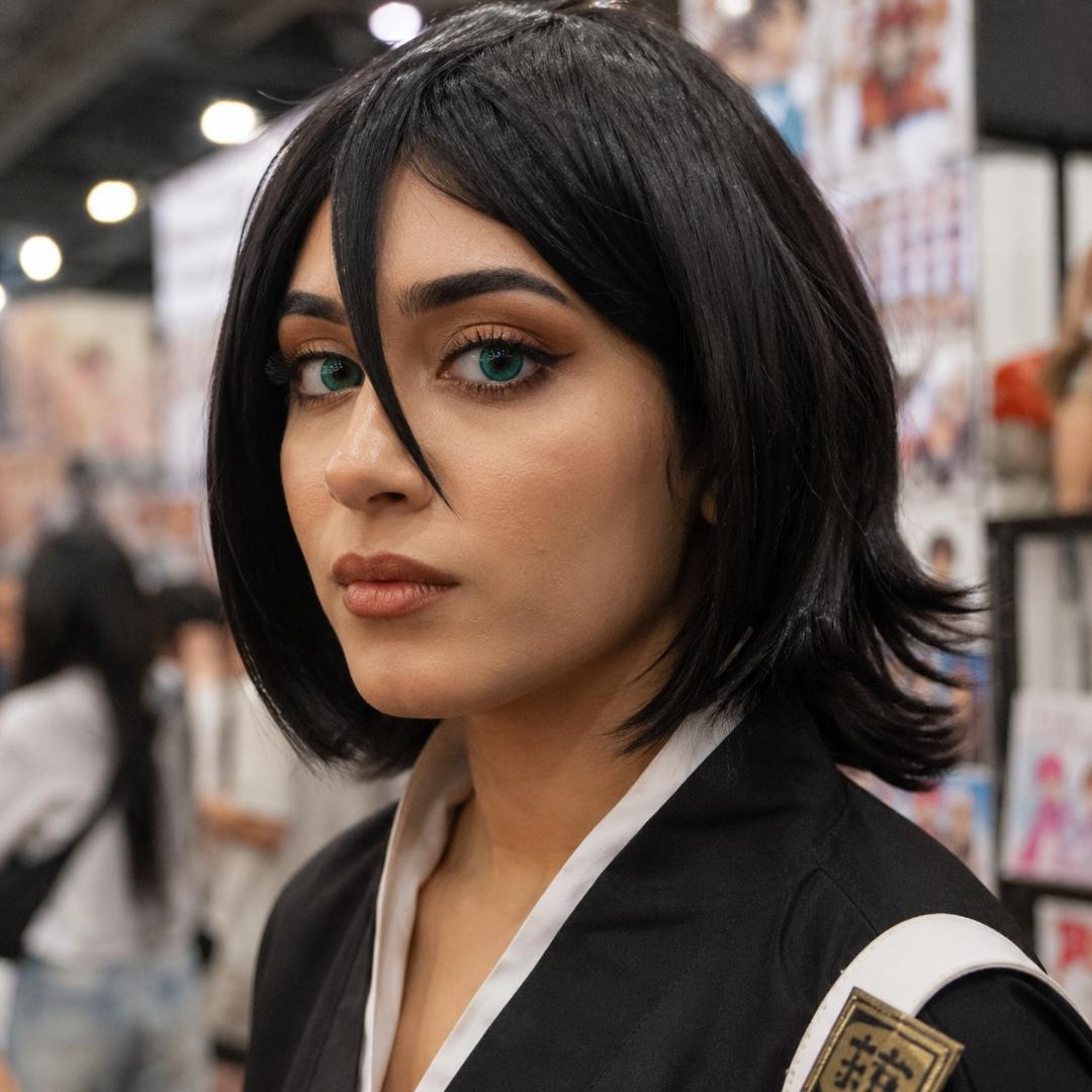 Convention portrait of a woman with a short black wig and cosplay outfit standing among blurred displays and attendees. Her serious expression and vivid green eyes give the character styling a dramatic effect.