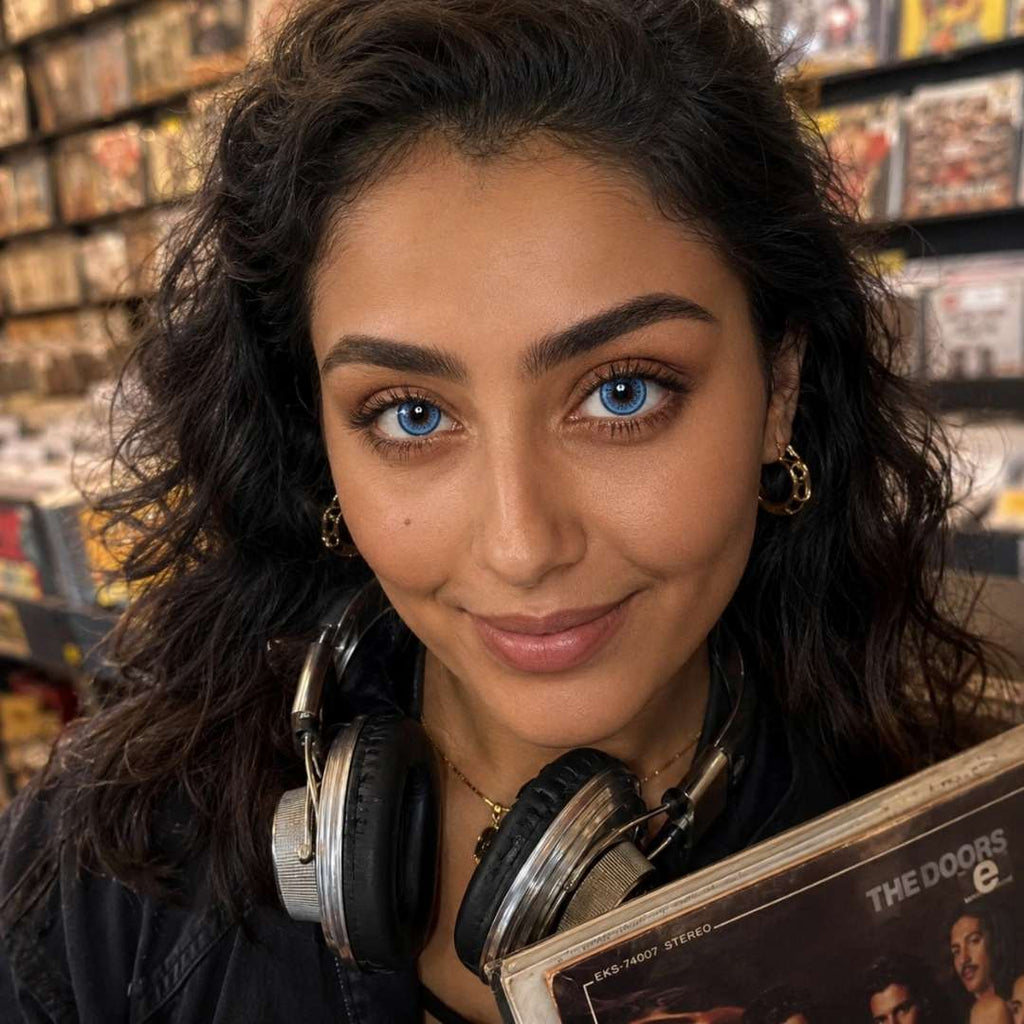 Smiling woman with curly dark hair wearing headphones around her neck while holding a vinyl record in a music store. Shelves of records fill the background and her vivid blue eyes stand out in the warm lighting.