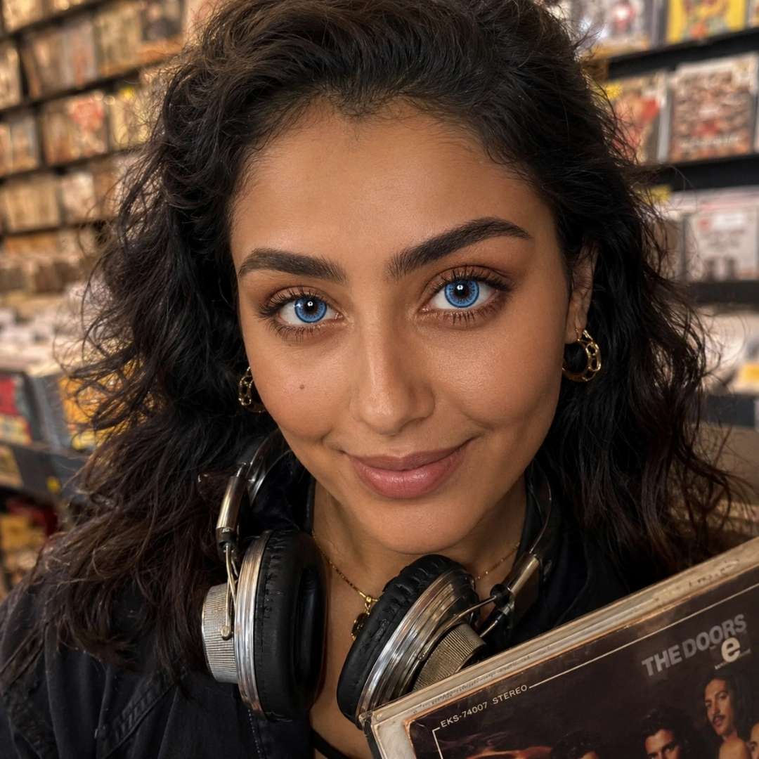 Smiling woman with curly dark hair wearing headphones around her neck while holding a vinyl record in a music store. Shelves of records fill the background and her vivid blue eyes stand out in the warm lighting.