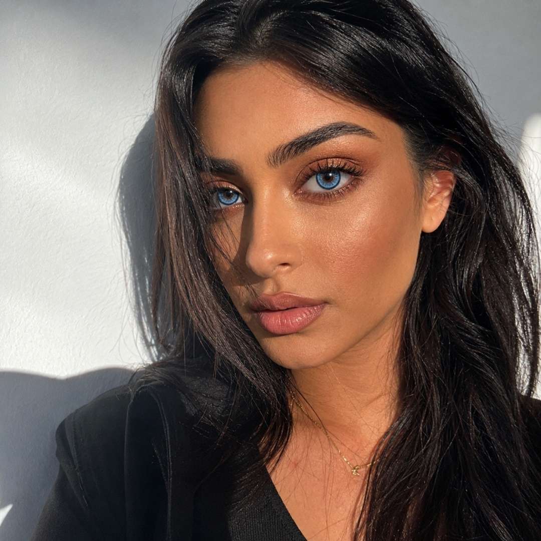 Close portrait of a woman with long dark hair and neutral makeup lit by angled sunlight against a plain wall. The strong shadows and vivid blue eyes create a dramatic mood.