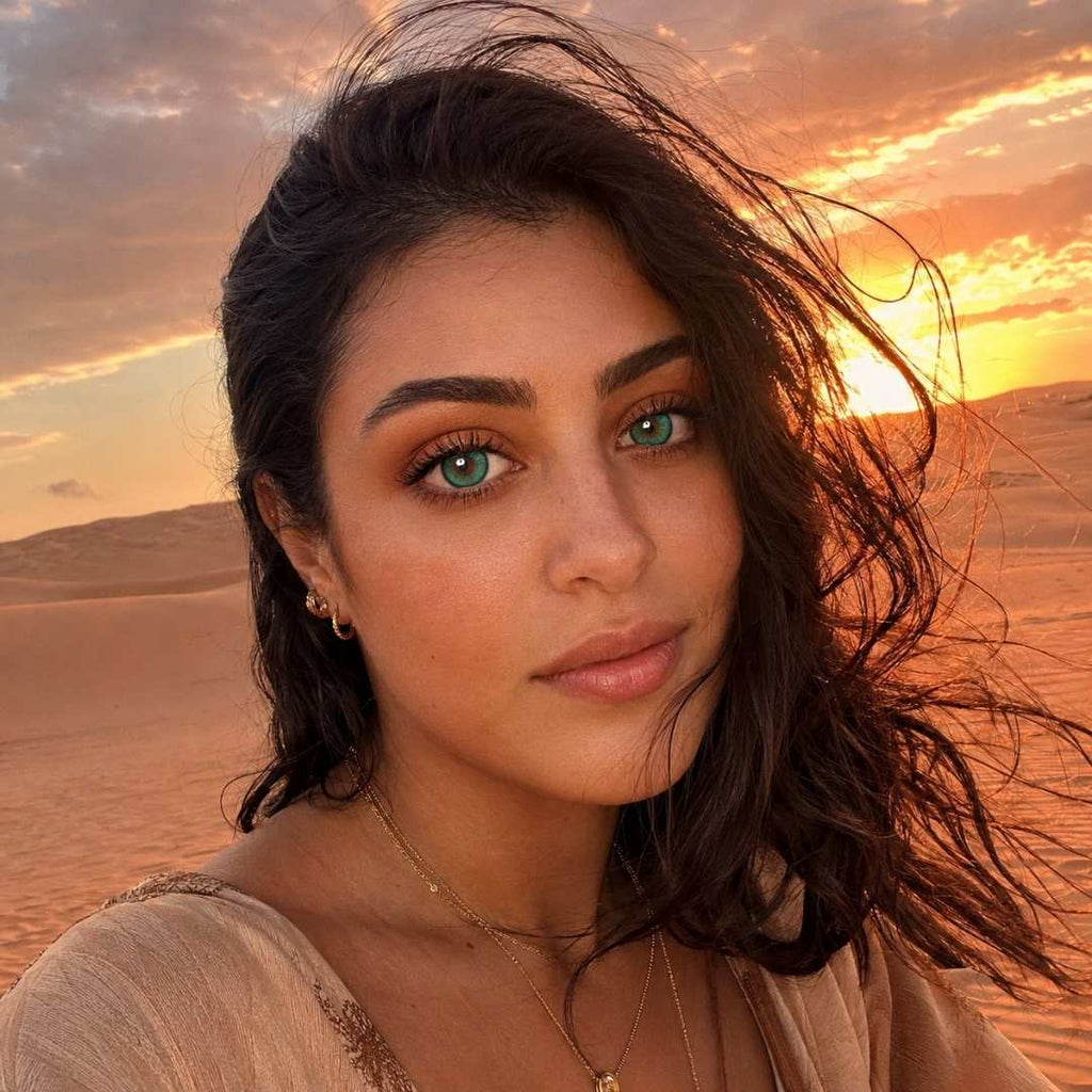 Woman with dark hair and dramatic green eyes photographed in a desert landscape at sunset. Warm golden light and blowing hair create a soft and cinematic mood.