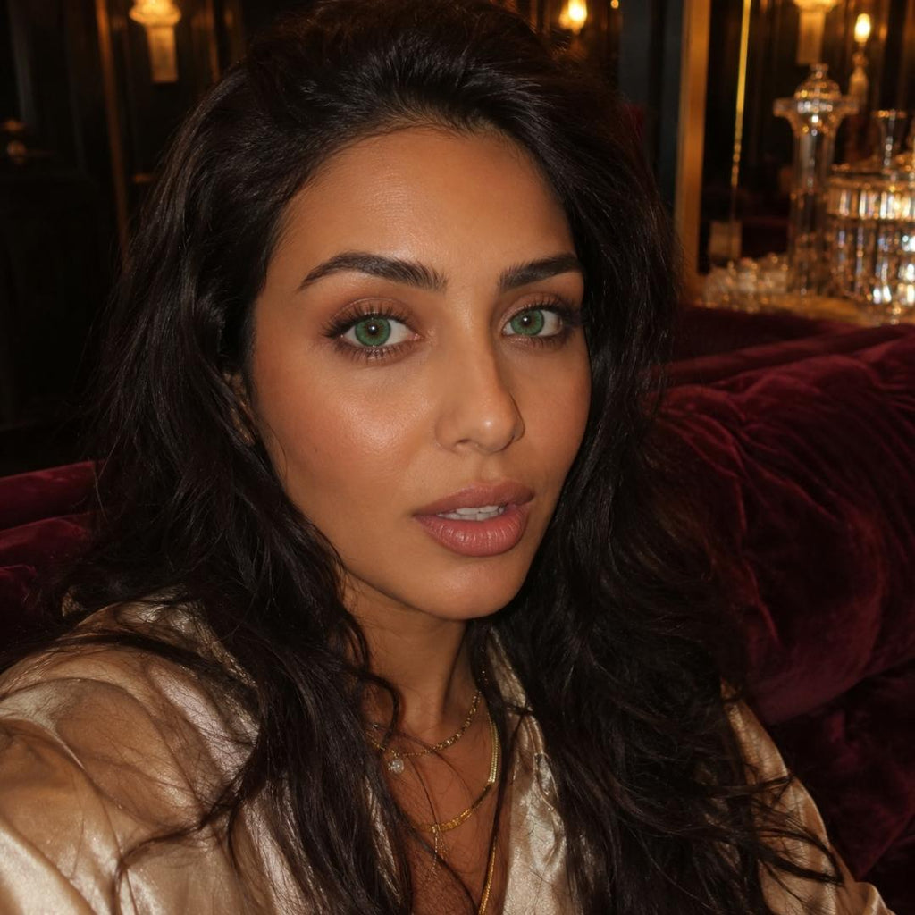 Indoor portrait of a woman with dark hair and green eyes seated against deep red velvet seating with warm lighting behind her. Gold jewelry and soft makeup add an elegant evening feel.