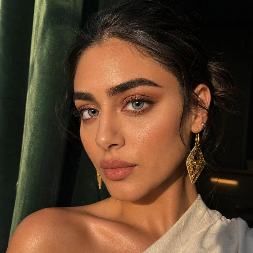 Close portrait of a woman with slicked back dark hair and gold geometric earrings in warm sunlight. Her striking gray eyes stand out against soft neutral makeup and a dark background.