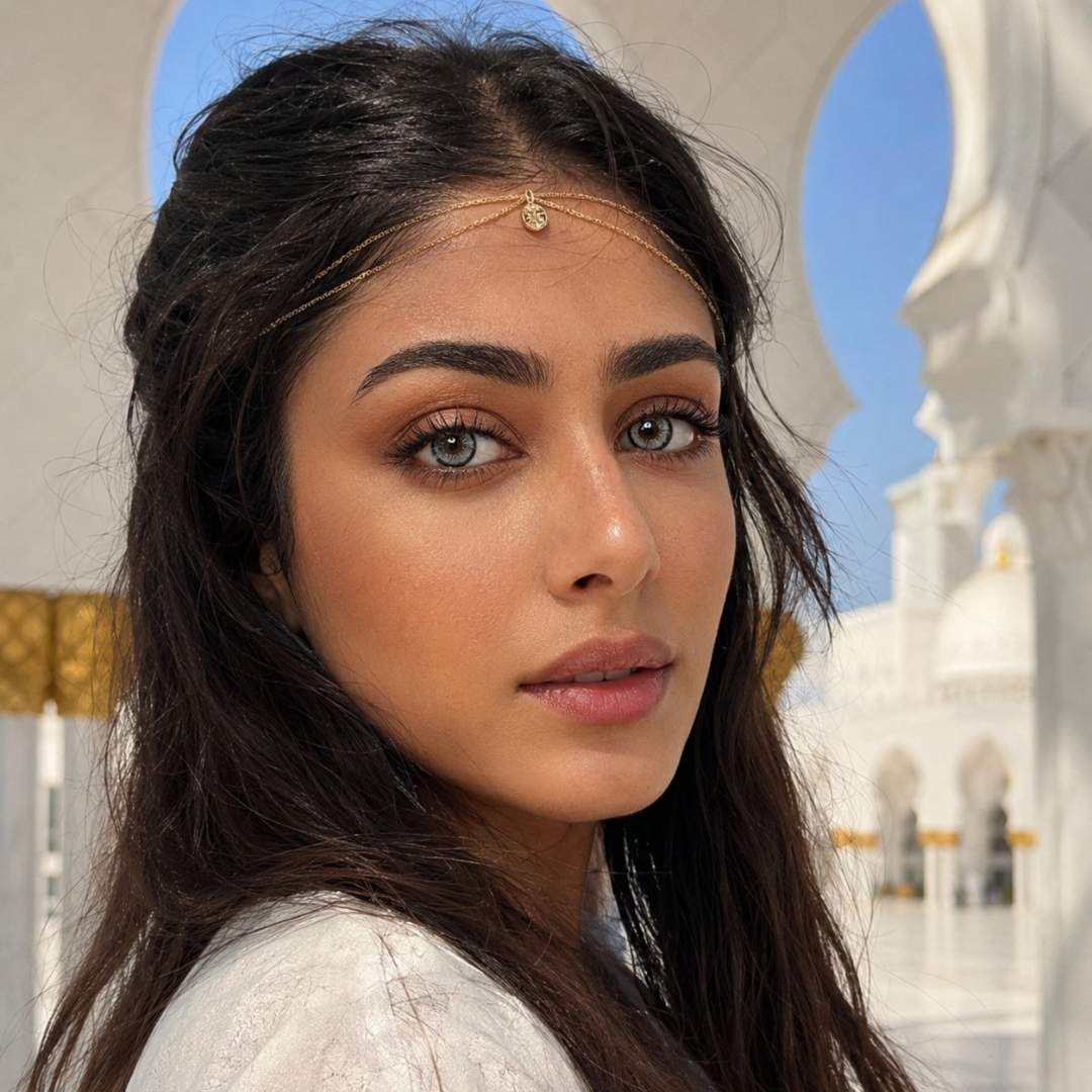 Woman with long dark hair and a delicate gold head chain stands beneath white arches with blue sky behind her. Her bold gray contacts and natural makeup create a soft elegant look.