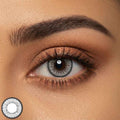 Close up of a single eye wearing a gray contact lens with a detailed patterned rim. Long lashes and warm brown eyeshadow frame the eye to highlight the lens color and texture.