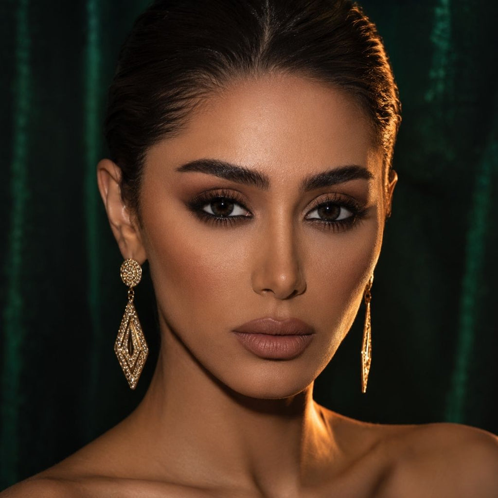 Glamour portrait of a woman with sleek pulled back hair and dramatic makeup wearing dark contact lenses. Gold statement earrings and moody lighting create an elegant and striking look.