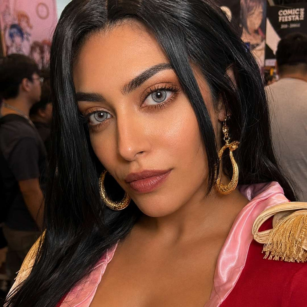 Portrait of a woman with long black hair and oversized gold earrings at a busy indoor event. Her gray eyes and soft glam makeup draw attention against a pink and red outfit.