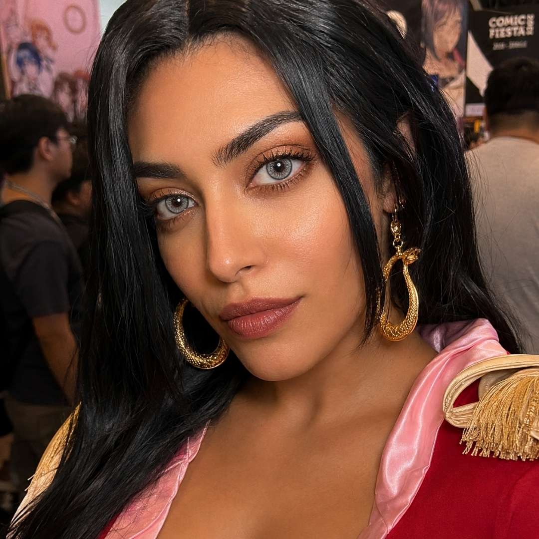 Portrait of a woman with long black hair and oversized gold earrings at a busy indoor event. Her gray eyes and soft glam makeup draw attention against a pink and red outfit.