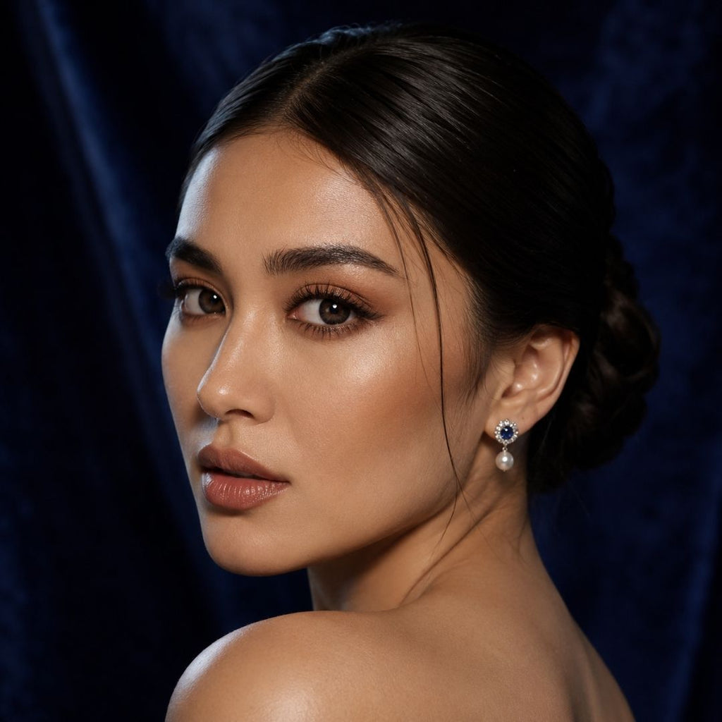 Side profile beauty portrait of a woman with hair styled in a low updo wearing dark contact lenses and pearl earrings. Soft glowing skin and deep blue background draw attention to the eyes and refined styling.