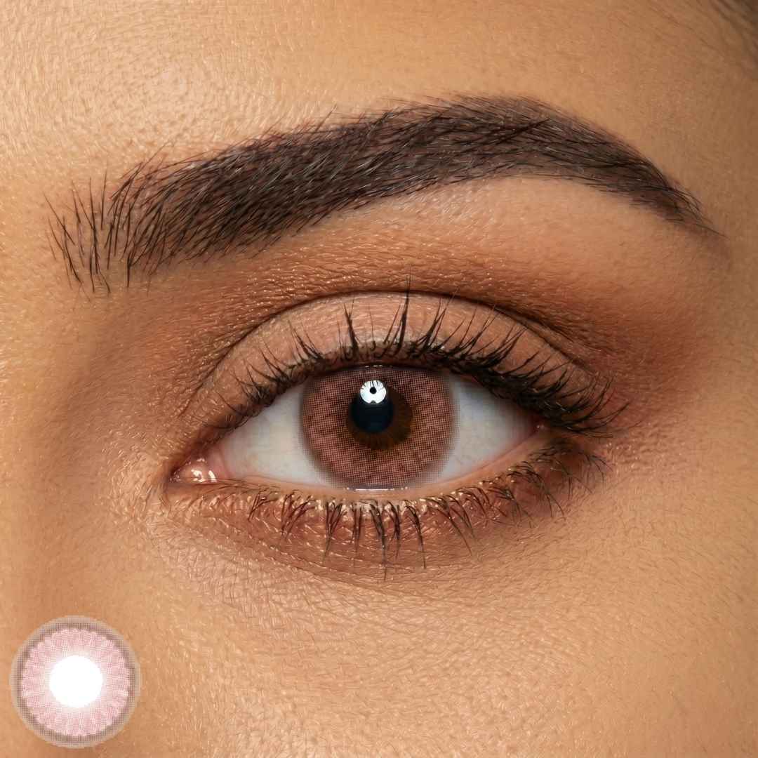 A close-up photograph focuses sharply on a woman's brown eye wearing a decorative, patterned pink-colored contact lens. Natural, groomed eyebrow, dark brown mascara, and soft copper-shimmer eyeshadow complement the distinct lens pattern. A standalone sample of the contact lens is visible at the bottom left.