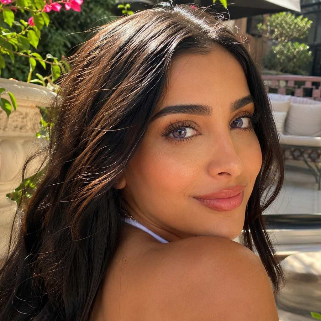 Woman with long dark hair and dreamy purple contact lenses looks back toward the camera while sitting outdoors in bright sunlight. Pink flowers and patio seating create a warm relaxed background setting.