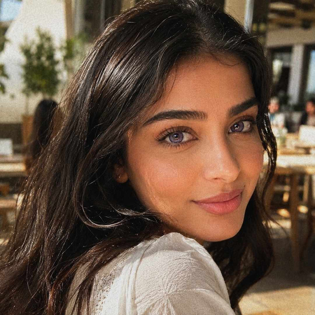 Close up portrait of a woman wearing dreamy purple contact lenses and a white textured blouse in a softly lit café. Golden sunlight casts warm shadows across her face and dark wavy hair.