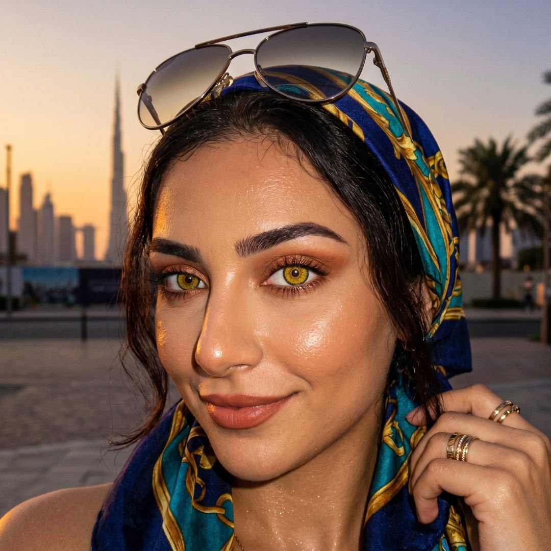Woman wearing a colorful headscarf with sunglasses on her head, standing in an urban setting with palm trees and buildings.