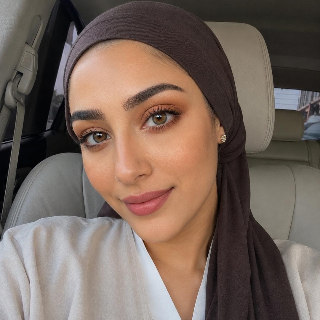 Woman wearing natural cacao colored lenses and a dark brown hijab takes a selfie while seated inside a car. Soft natural light highlights her warm makeup tones and glossy pink lips against the cream leather interior.