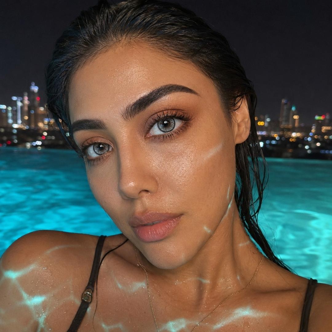 Nighttime poolside selfie with city lights glowing in the background and water reflections across the skin. Wet dark hair and striking gray eyes create a dramatic look.