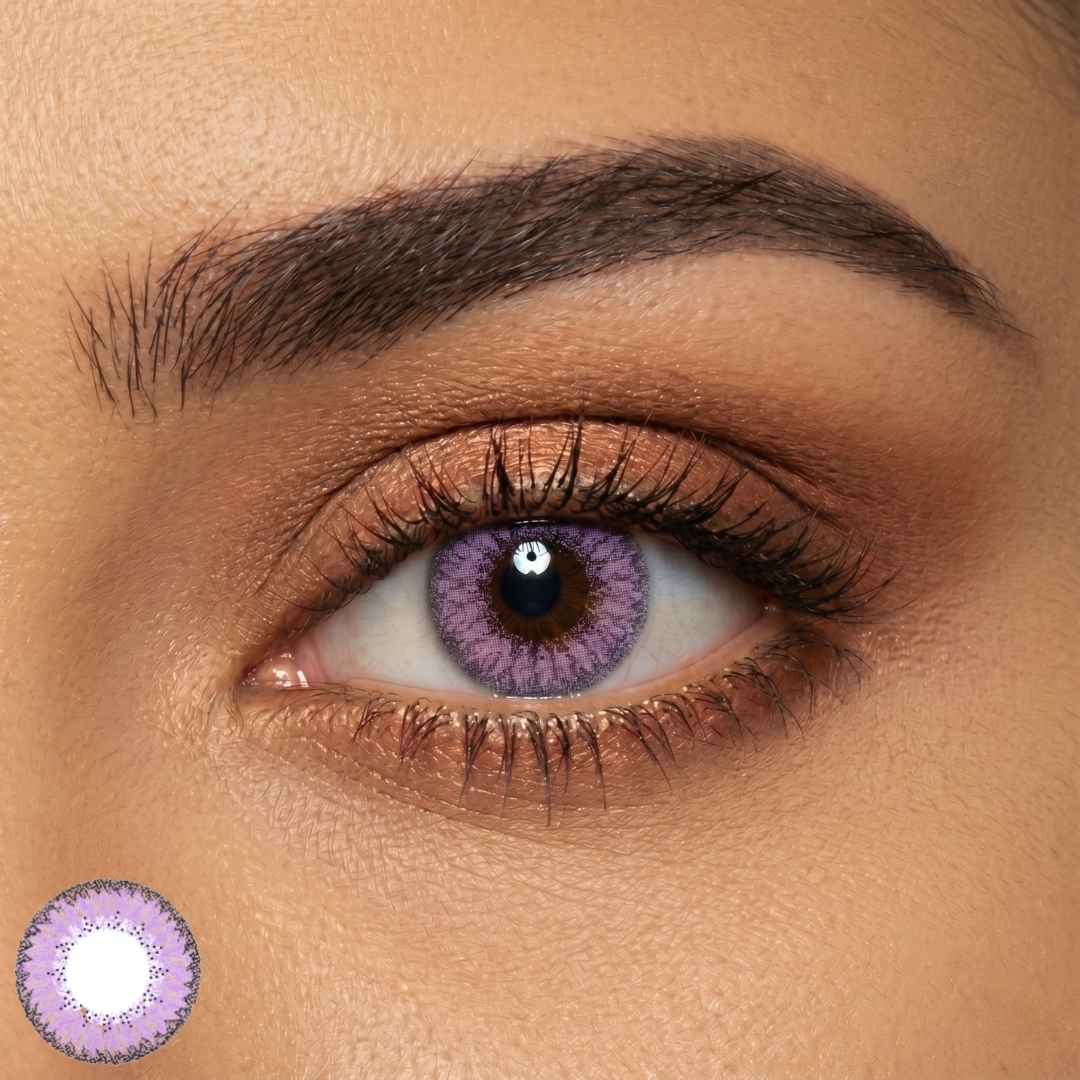 Detailed close up of an eye wearing dreamy purple contact lenses with a soft lavender tone and darker violet outer ring. Shimmery bronze eyeshadow and long lashes enhance the vibrant lens color.