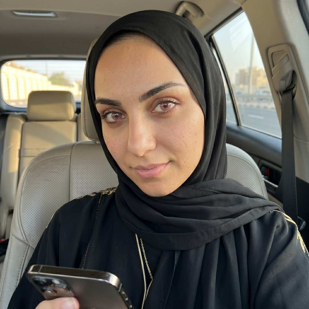 A candid portrait captures a young woman with warm brown eyes, wearing a modest black hijab and abaya with gold embroidery, seated as a passenger in a luxury car in Dubai. Her gaze is confident, holding an iPhone with a triple-lens camera. The soft daylight and blurred city background complete the high-end look.