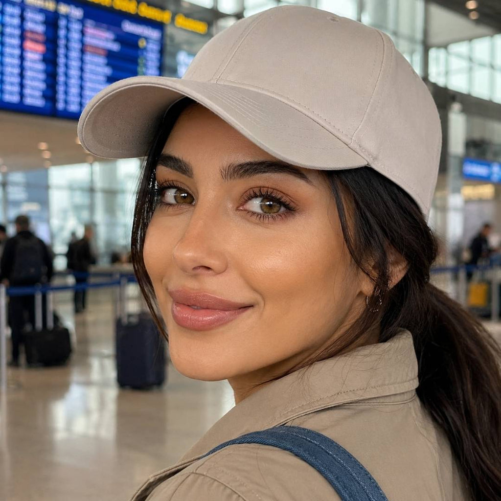 Woman wearing natural looking brown colored contact lenses and a beige baseball cap turns back toward the camera inside an airport terminal. Blurred flight information boards and travelers with luggage appear in the background.