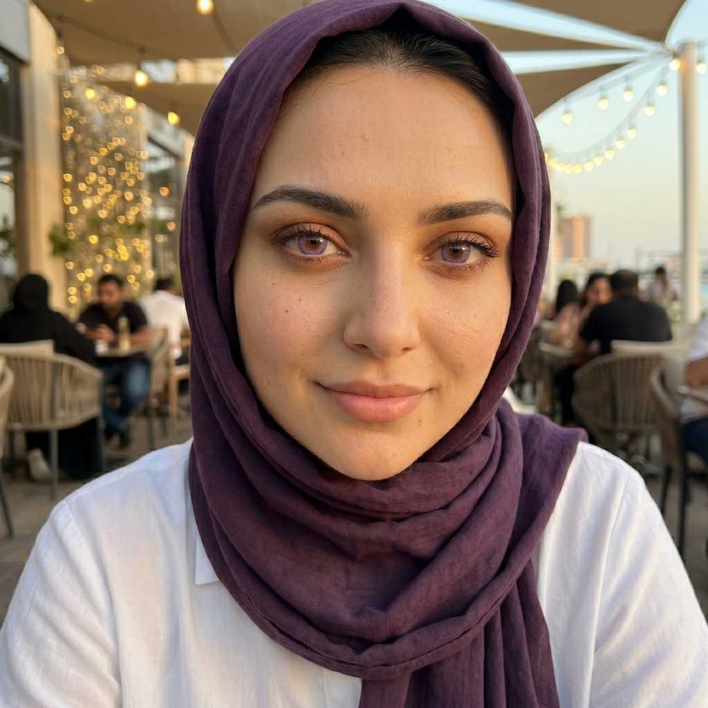 Woman wearing a purple hijab and dreamy purple contact lenses sits at an outdoor café with string lights glowing in the background. Soft evening light highlights her natural freckles and subtle makeup.