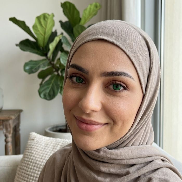 Woman wearing a beige hijab in a room with plants and a window.