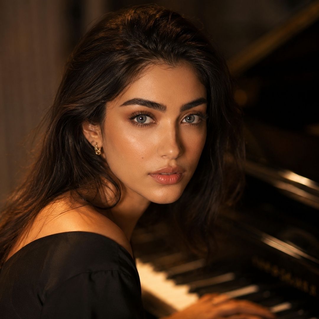 Woman with dark wavy hair and soft gray eyes looks toward the camera while seated beside a piano. She wears a black off shoulder top in a dimly lit setting with blurred piano keys in the background.