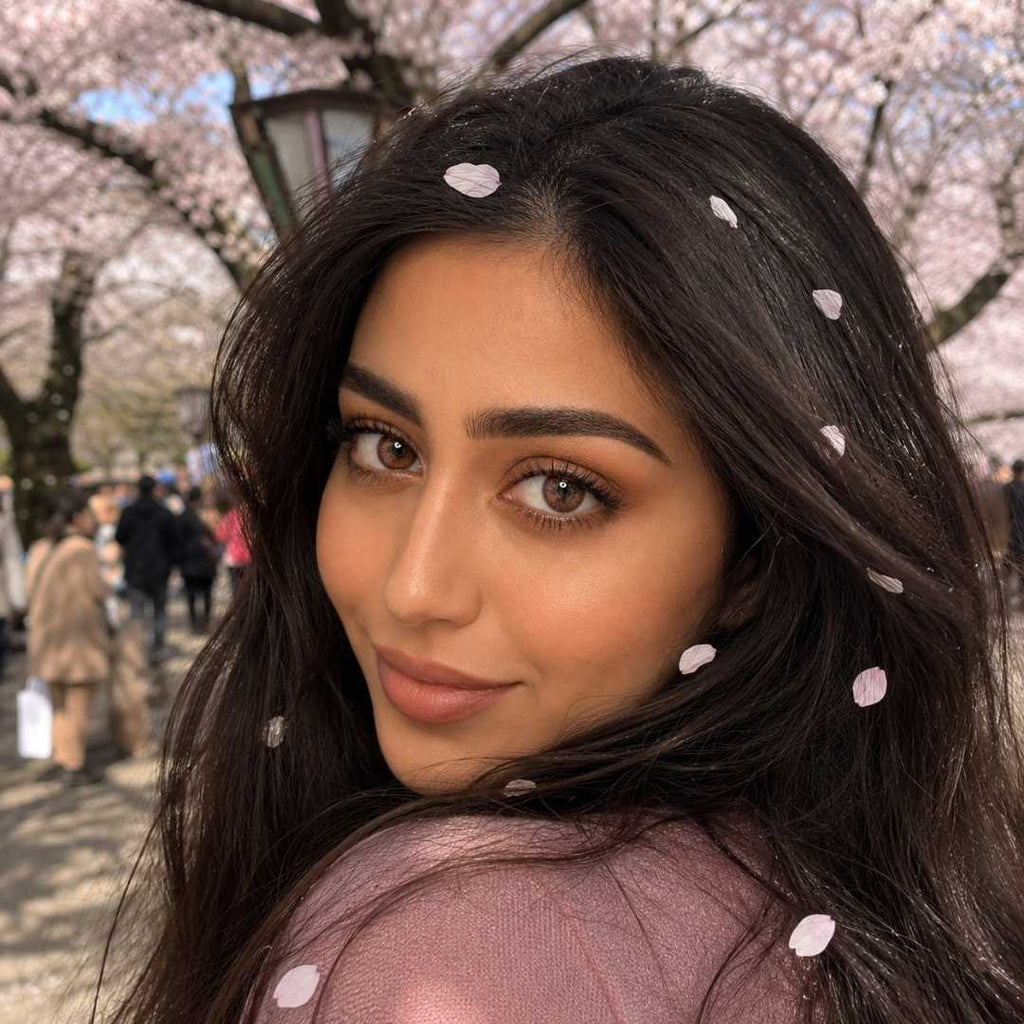 A portrait photograph of a woman with long dark hair, looking back over her shoulder with a playful smile. Her hair and shoulder are lightly covered with fallen pink cherry blossom petals. The background is a soft-focus (bokeh) view of a path lined with flowering cherry trees (sakura) in full bloom on a sunny day
