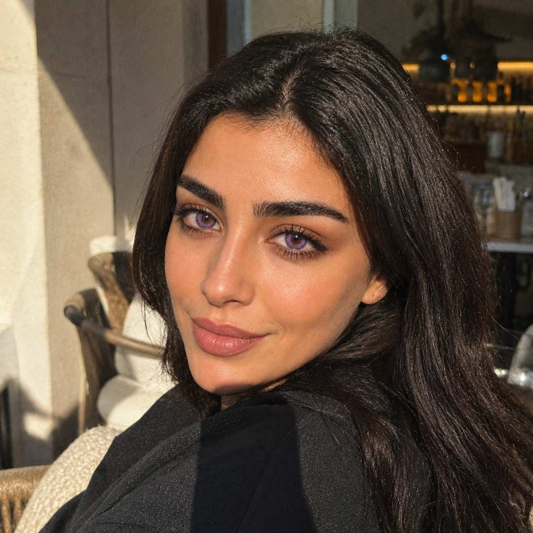 Woman with long dark hair and subtle violet color contacts sits outdoors in warm afternoon sunlight. Soft shadows and café seating in the background create a relaxed and elegant atmosphere.
