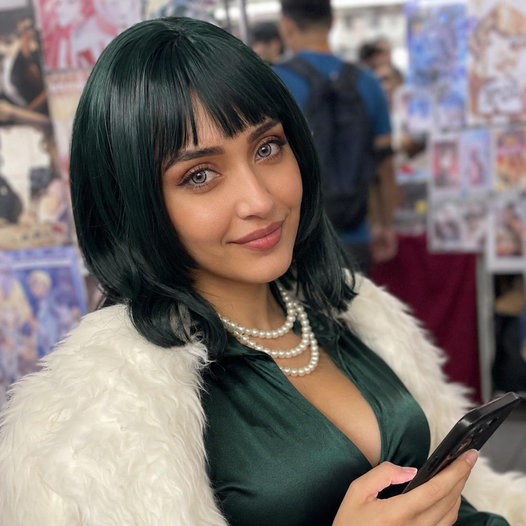 Woman wearing a dark green wig and pearl necklace smiles while holding a phone at a convention. Her gray eyes and vintage inspired outfit create a polished cosplay look.