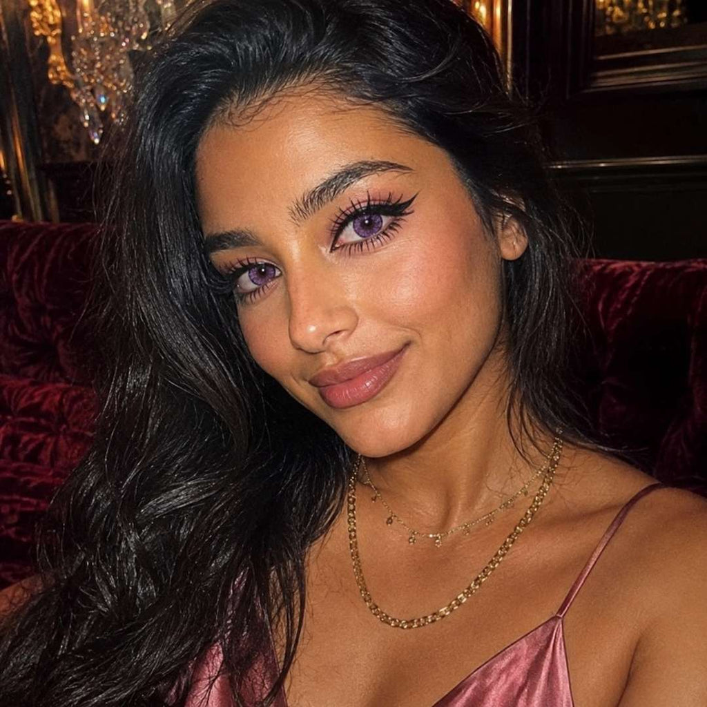 Evening portrait with long dark hair, layered gold necklaces, and soft pink colored contacts. Warm ambient lighting enhances the subtle pink hue of the lenses against naturally dark eyes.