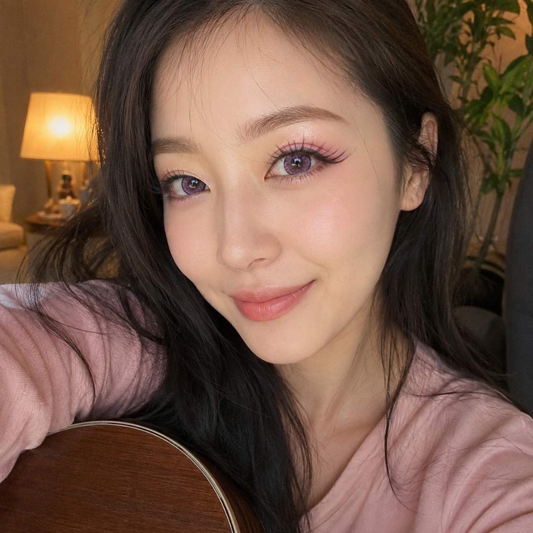 Indoor selfie of a person with long dark hair holding an acoustic guitar beside a warm table lamp. Soft pink colored contacts add a subtle pastel tone to the eyes while keeping a natural appearance.
