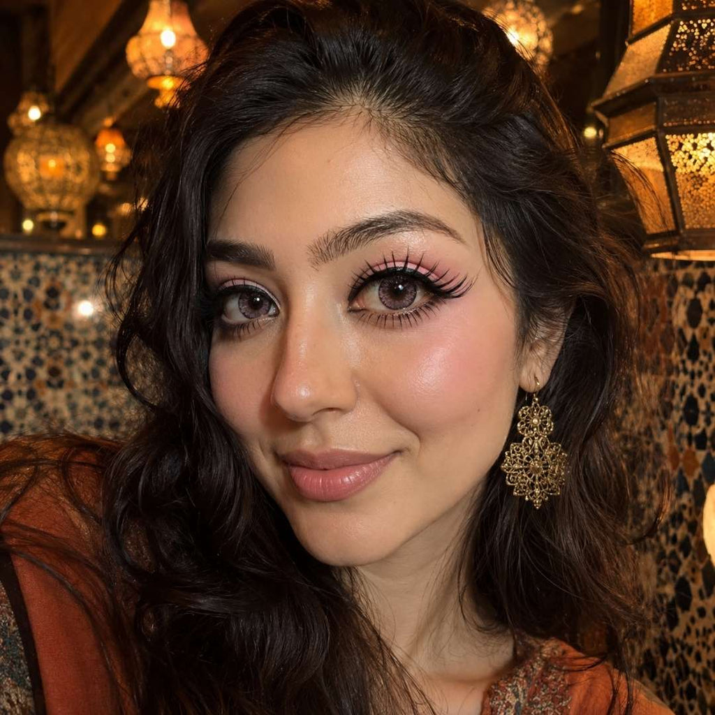 Portrait in a warmly lit restaurant featuring dark wavy hair, ornate earrings, and kawaii contact lenses in a soft pink shade. Decorative lanterns and patterned walls frame the scene.