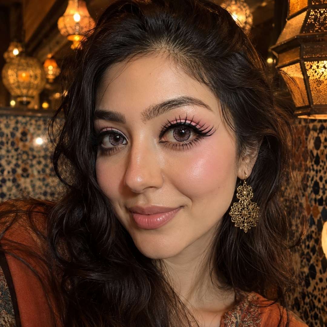 Portrait in a warmly lit restaurant featuring dark wavy hair, ornate earrings, and kawaii contact lenses in a soft pink shade. Decorative lanterns and patterned walls frame the scene.