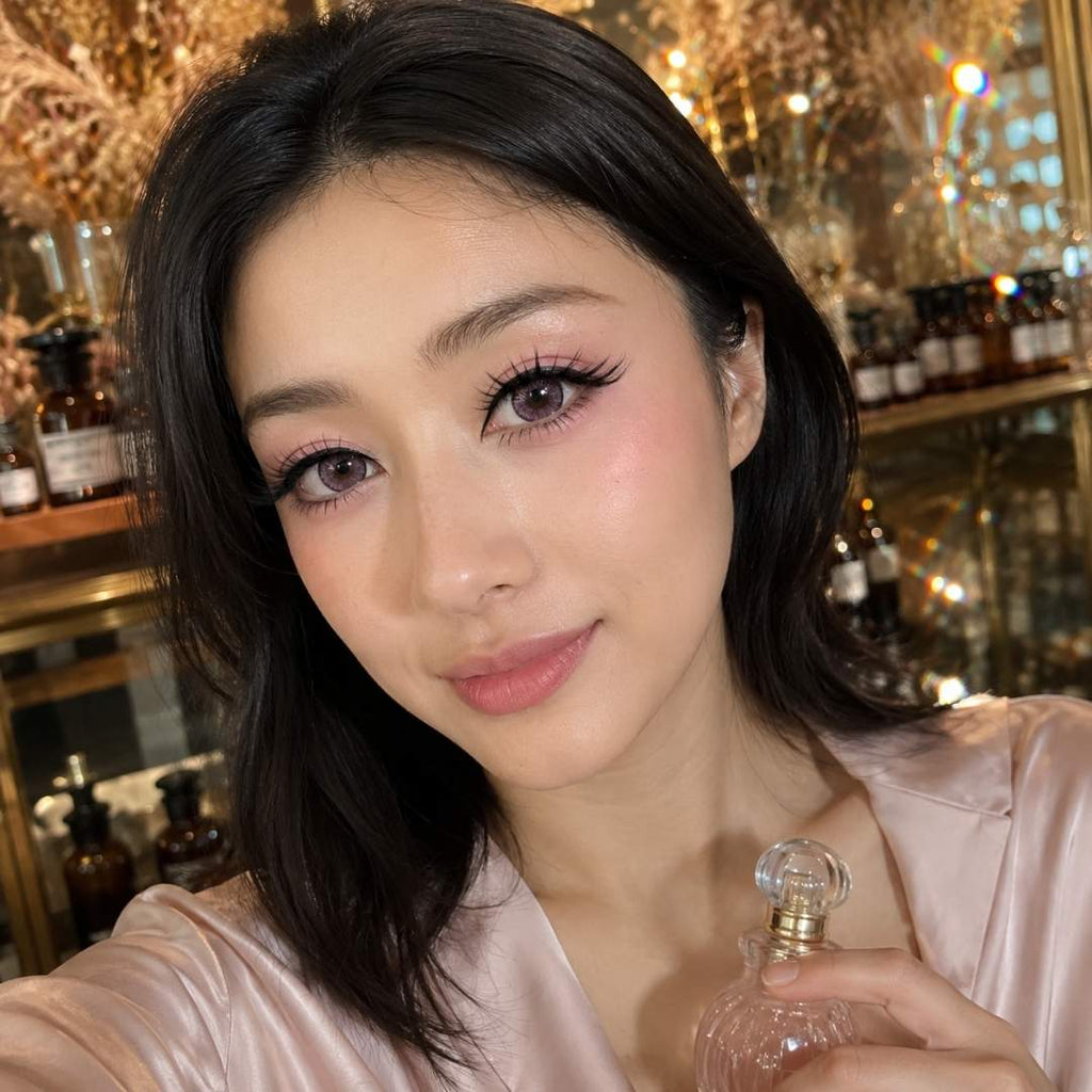Beauty portrait in a boutique style setting with a glass perfume bottle held in the foreground. Pink contact lenses complement the soft makeup and warm golden lighting.