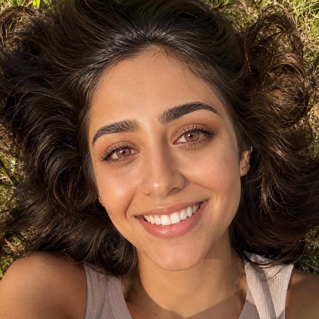 An overhead, selfie-style photograph of a woman with long, dark wavy hair spread out wildly around her head, lying comfortably in green grass on a sunny day. She is smiling broadly, showing her teeth, directly up at the camera. Warm, natural sunlight is on her face and the grass.