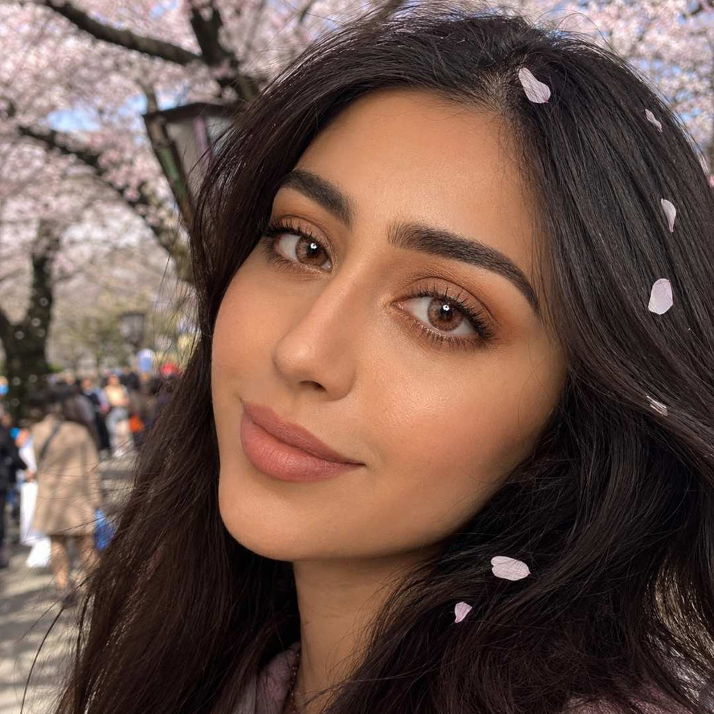 A close-up portrait of a woman with long, wavy dark brown hair, featuring small pink cherry blossom petals scattered through her hair. She is smiling softly at the camera. The background is a beautifully blurred scene of flowering cherry trees (sakura) in a park with distant, out-of-focus people