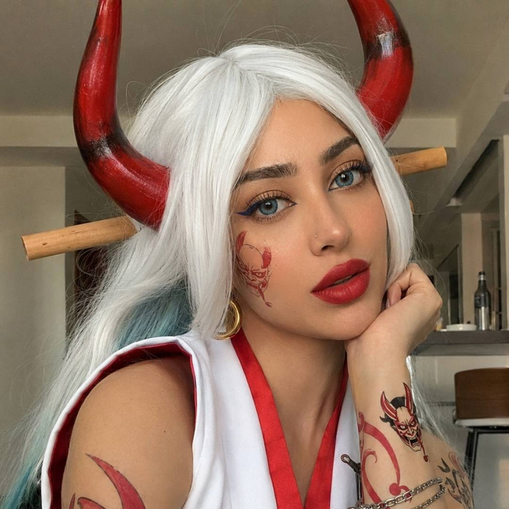 Woman wearing cosplay blue contacts, a white wig, and red horn accessories poses in a demon inspired cosplay look. Red face and arm markings with bold red lipstick add dramatic detail to the costume.